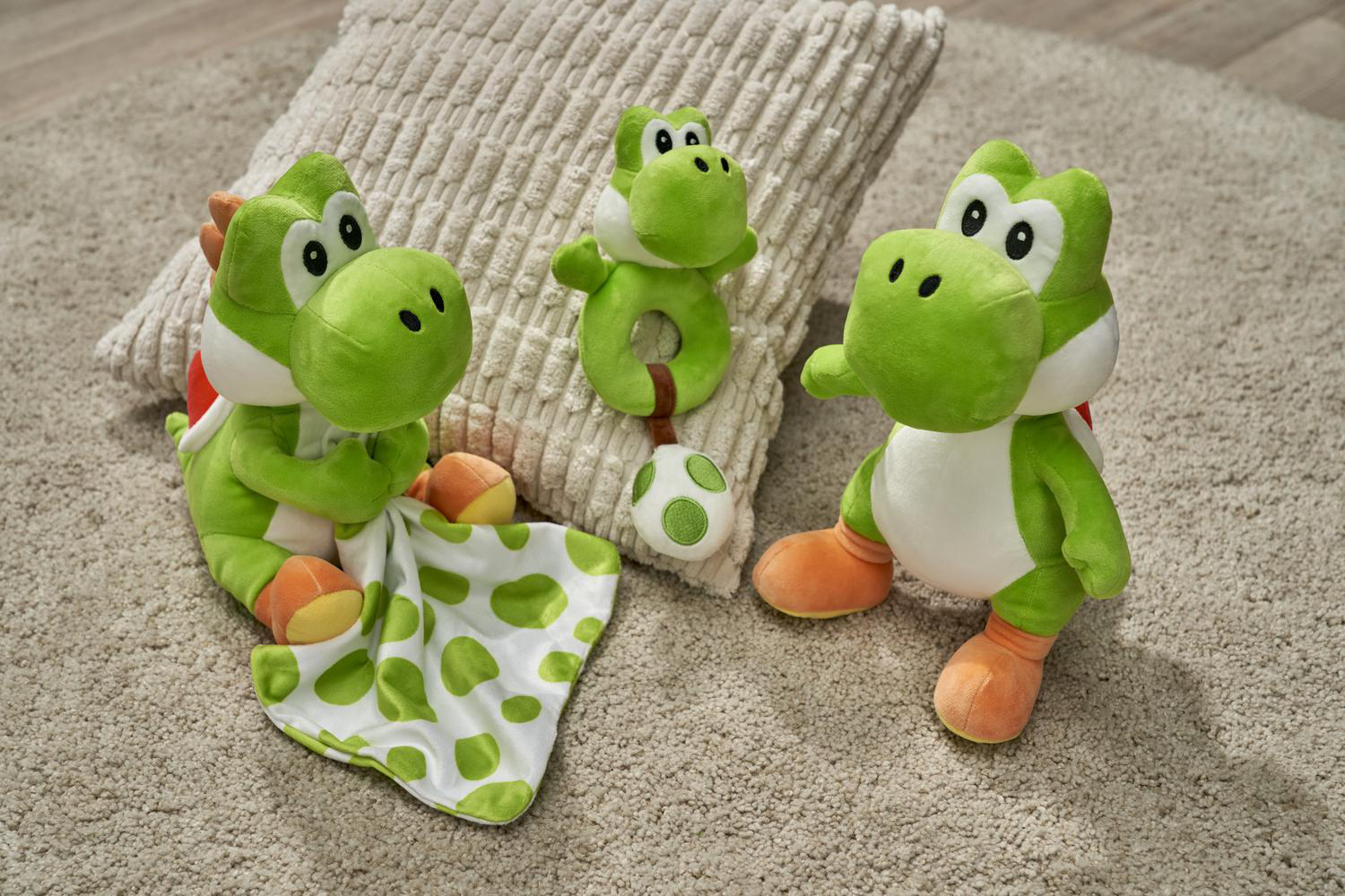 Drei grüne Yoshi-Plüschtiere liegen auf einem Teppich, eines mit einer Decke.