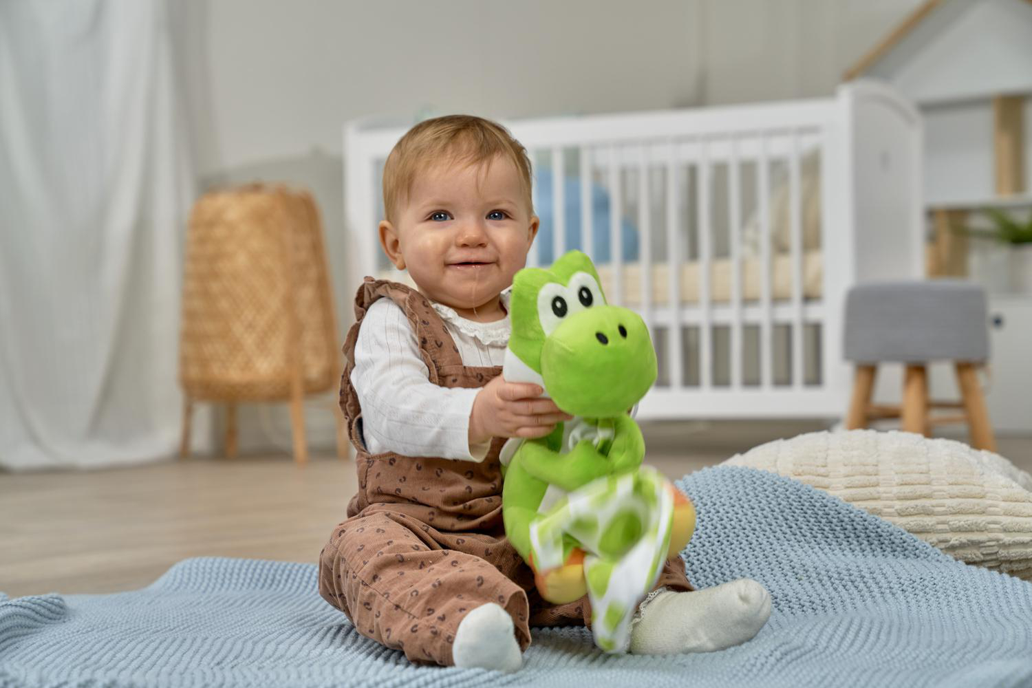 Ein Baby sitzt auf einer Decke und hält eine grüne Plüschfigur. Ein Kinderbett ist im Hintergrund.
