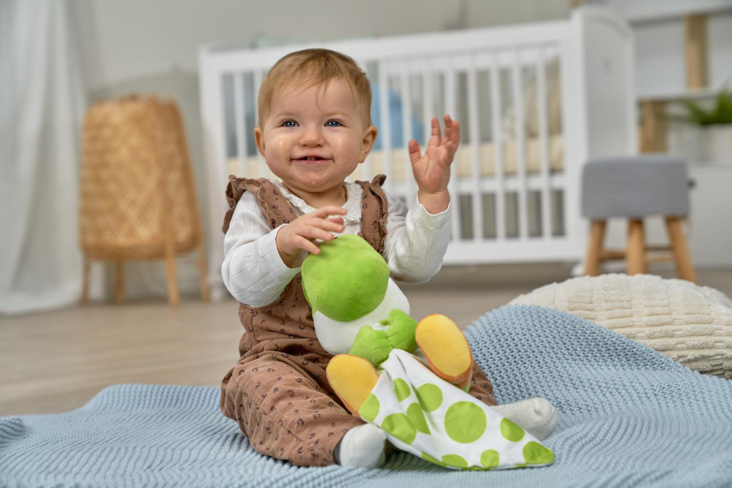 Ein Baby sitzt auf einer Decke und hält ein grünes Spielzeug. Ein Kinderbett ist im Hintergrund.