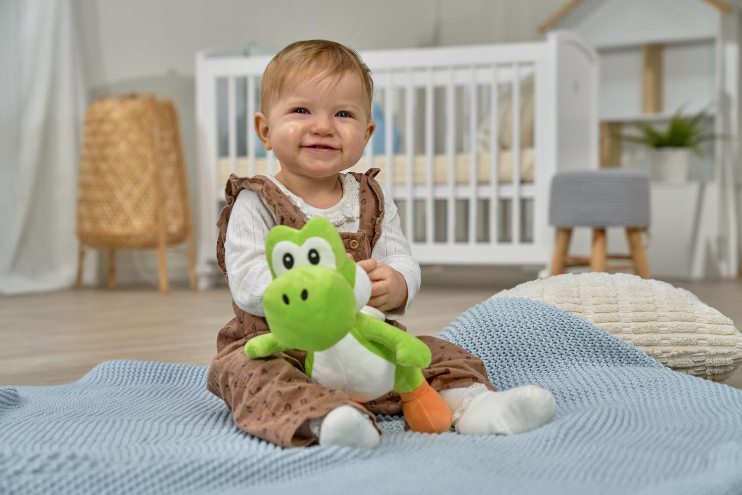 Ein Baby sitzt auf einer blauen Decke und hält eine grüne Yoshi-Puppe. Ein Kinderbett im Hintergrund.