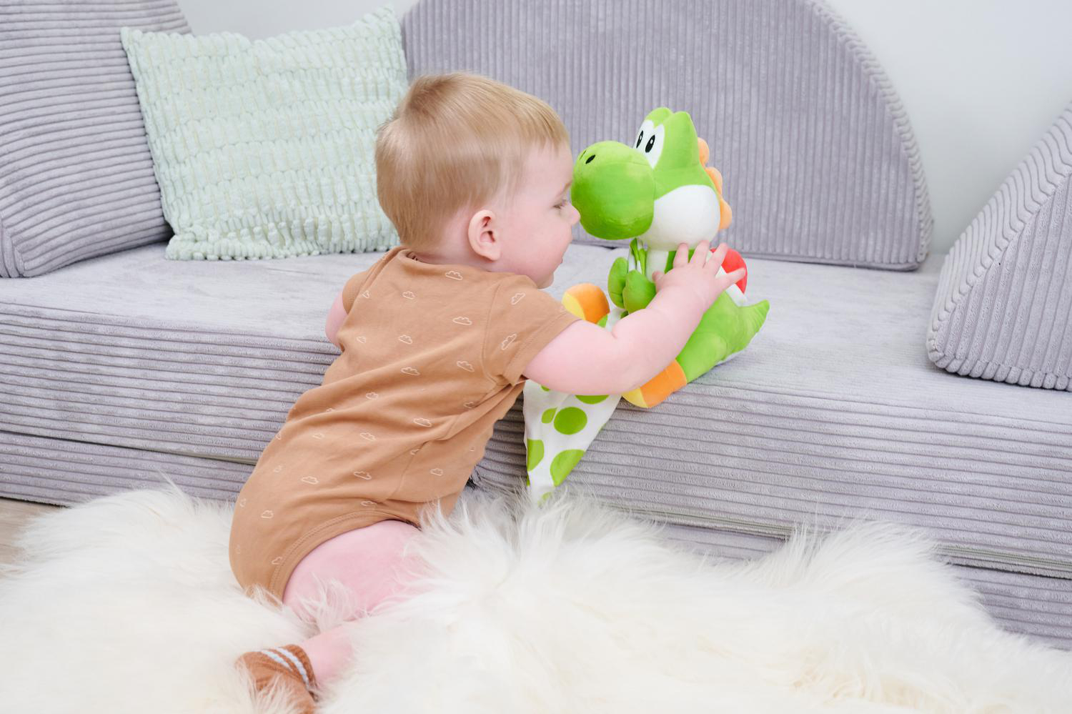 Ein Baby spielt mit einem grünen Yoshi-Plüschtier auf einem weißen Teppich.