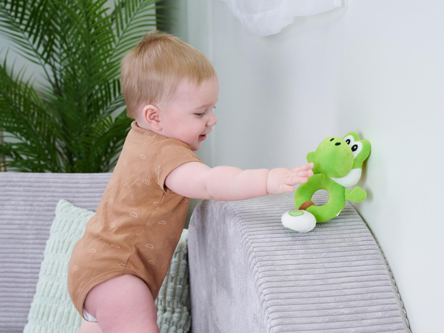 Ein Baby in einem braunen Strampler greift nach einem grünen Yoshi-Spielzeug an einer Wand.