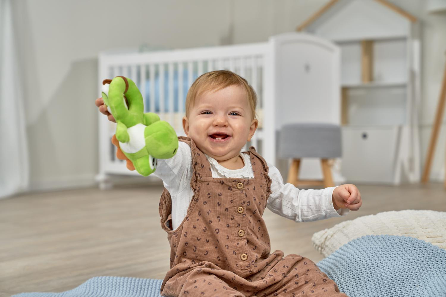 Ein Baby hält ein grünes Yoshi-Plüschtier, lächelt und sitzt auf Kissen.
