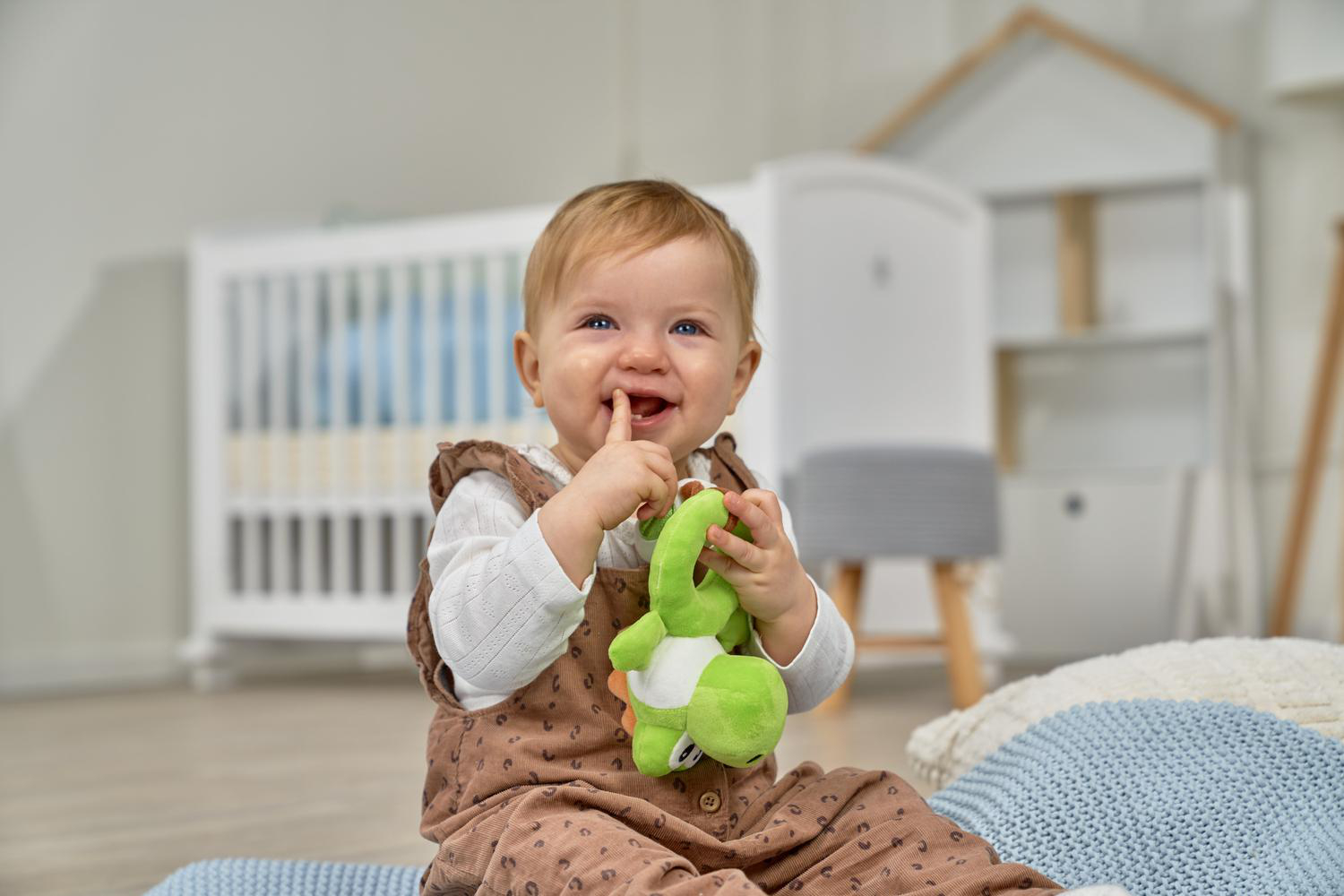 Ein lächelndes Baby sitzt und hält ein grünes Spielzeug. Ein Kinderbett ist im Hintergrund.