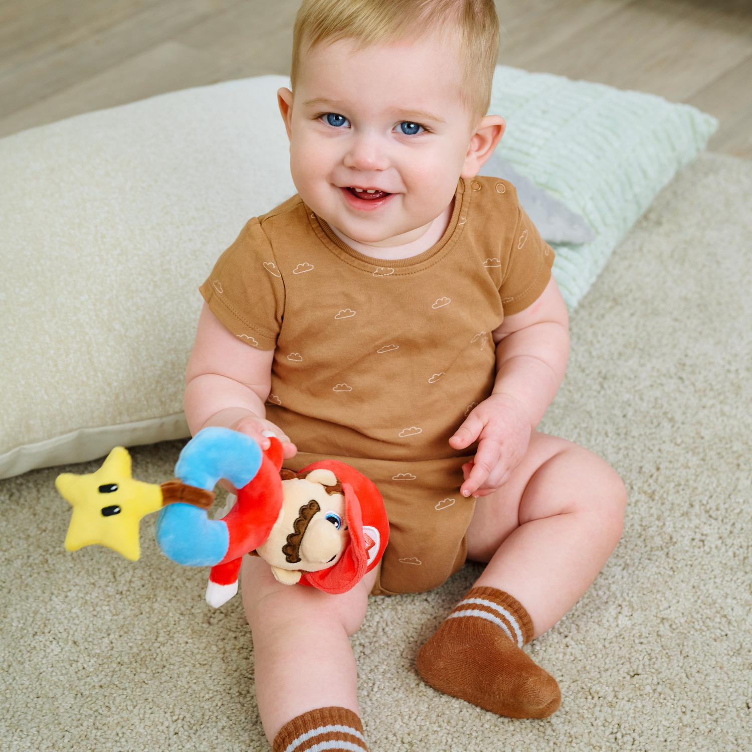 Ein Baby mit blauen Augen sitzt auf einem Teppich und hält ein Mario-Spielzeug.
