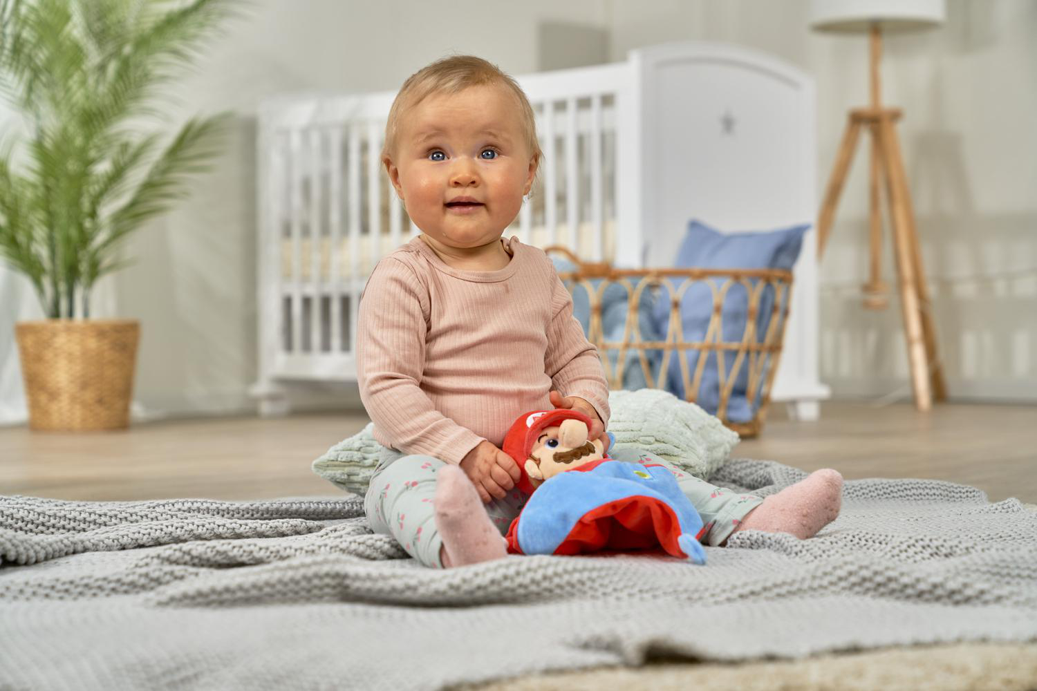 Ein Baby sitzt auf einer Decke und hält eine Mario-Puppe. Ein Kinderbett im Hintergrund.