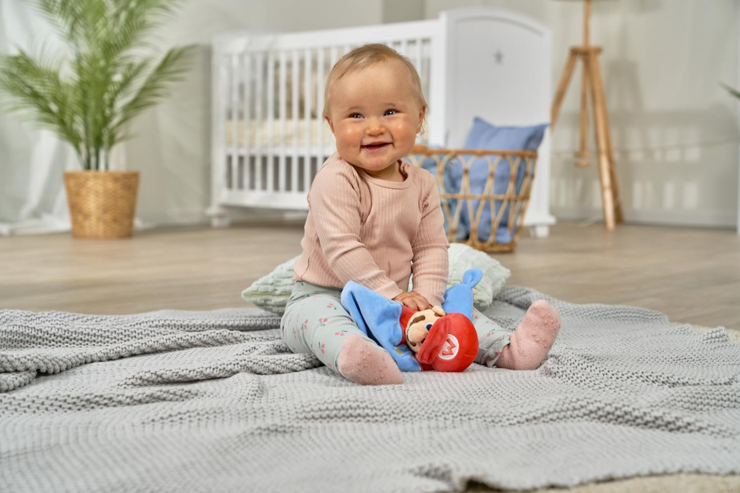 Ein Baby sitzt auf einer grauen Decke und hält ein Mario-Spielzeug.