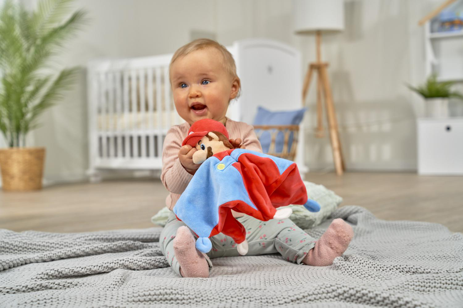 Ein Baby hält eine Mario-Puppe auf einer Decke vor einem Kinderbett.