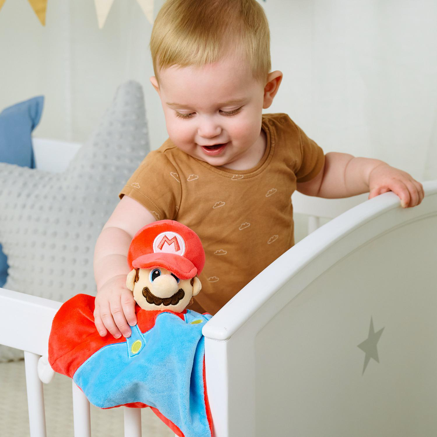 Ein Baby im Kinderbett hält ein Mario-Plüschtier. Das Baby trägt ein braunes Hemd mit Wolken.