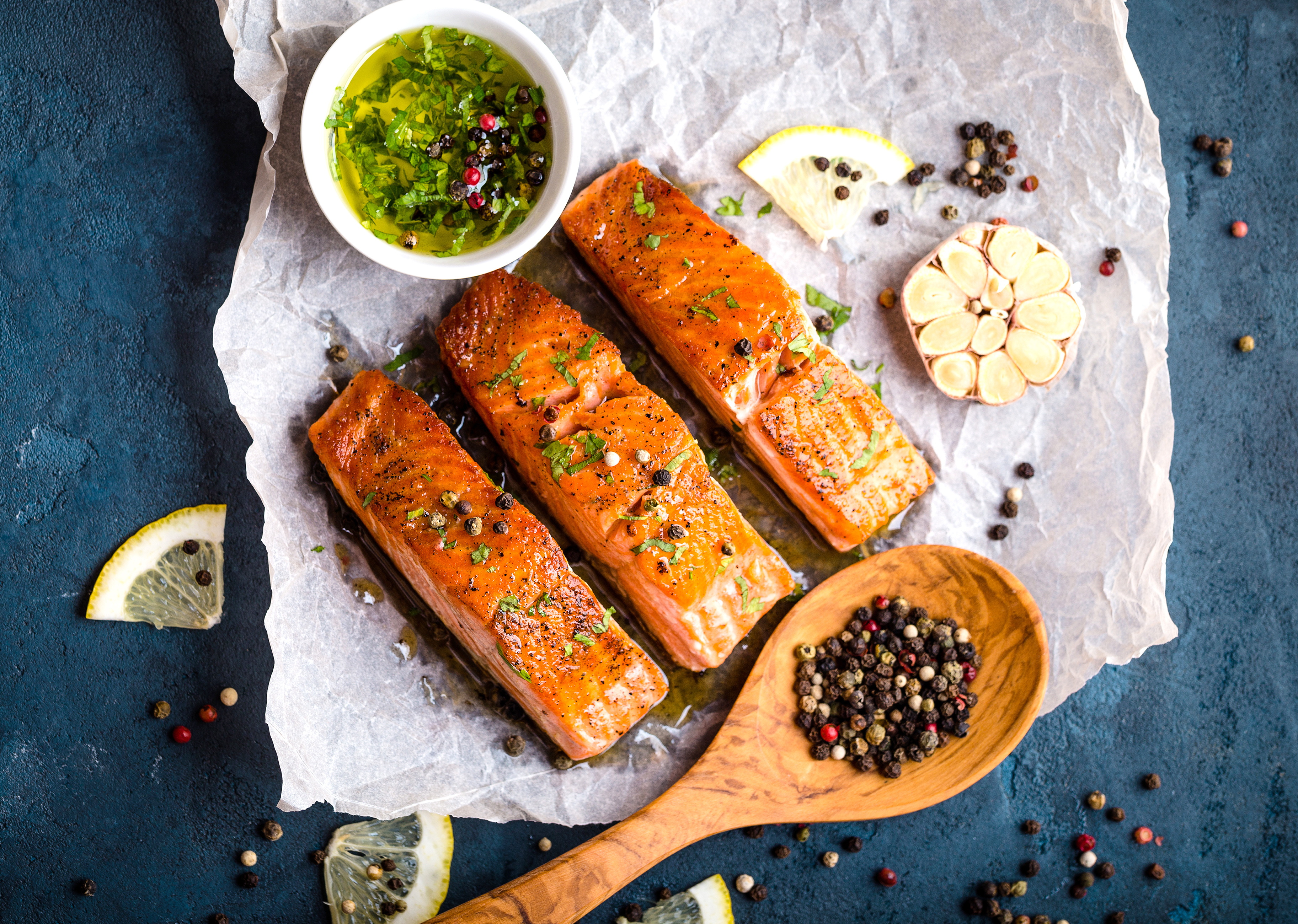 Drei gebratene Lachsfilets auf Backpapier mit Zitrone, Knoblauch und Pfefferkörnern.