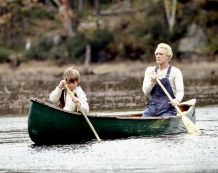 Ein Mann in Latzhosen und ein Junge rudern in einem grünen Kanu auf einem See.