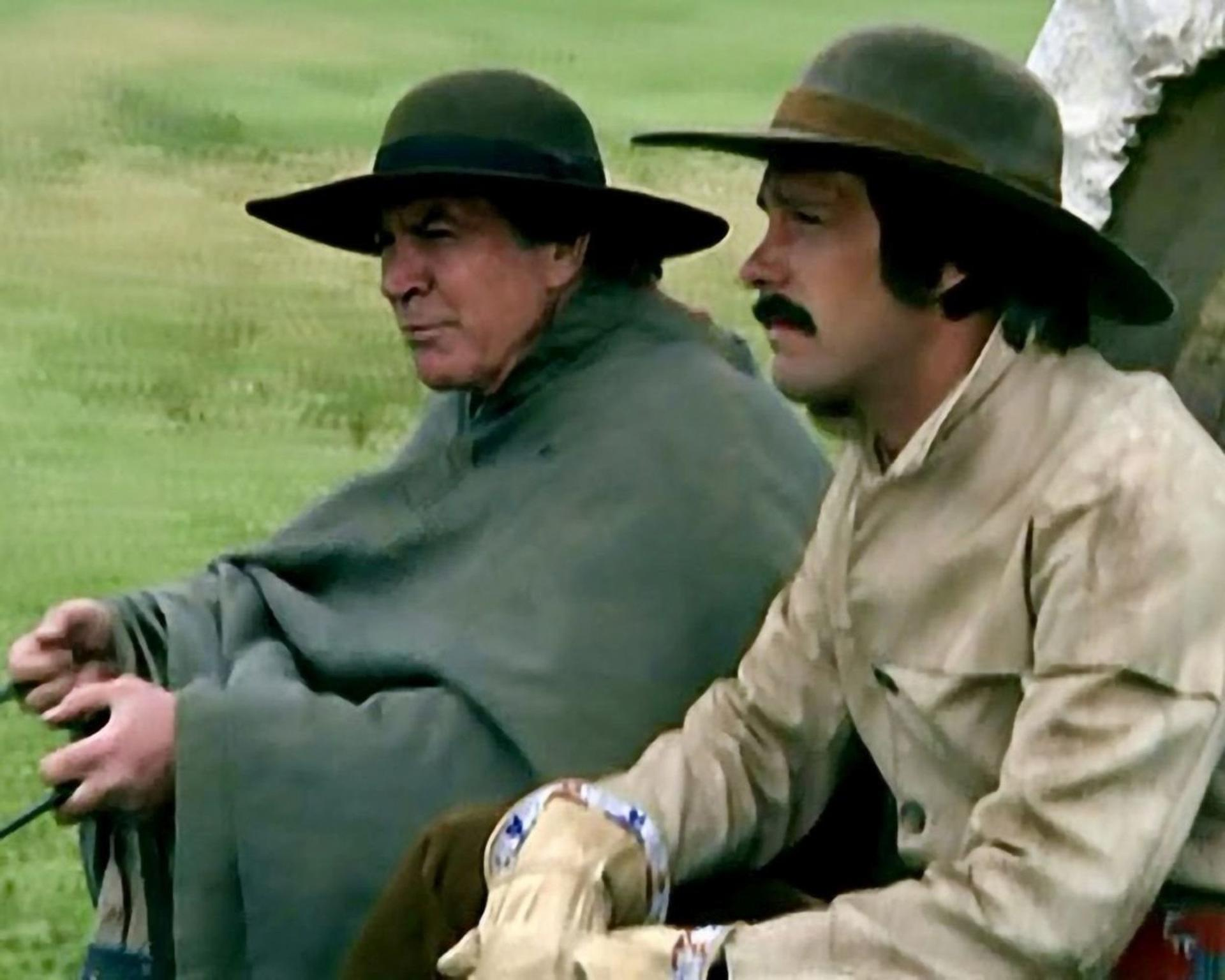 Zwei Männer in Hüten und Kleidung der Zeit sitzen in einem Wagen auf einem Feld.