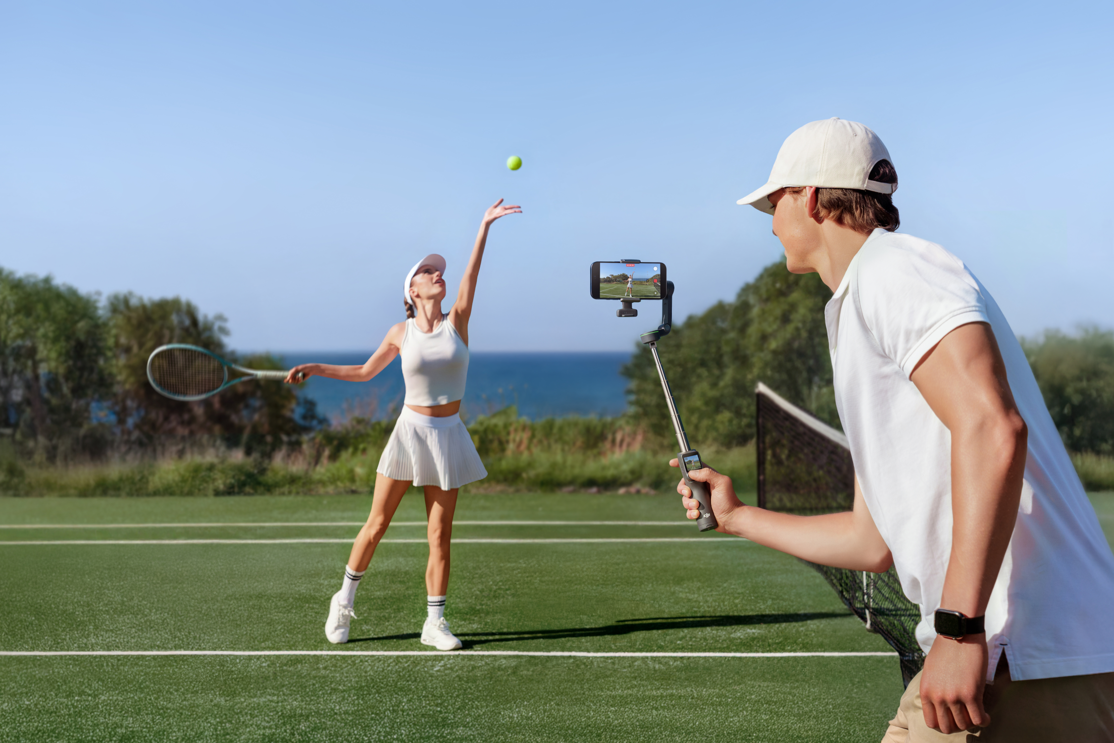 Un uomo filma una donna che serve una palla da tennis su un campo.