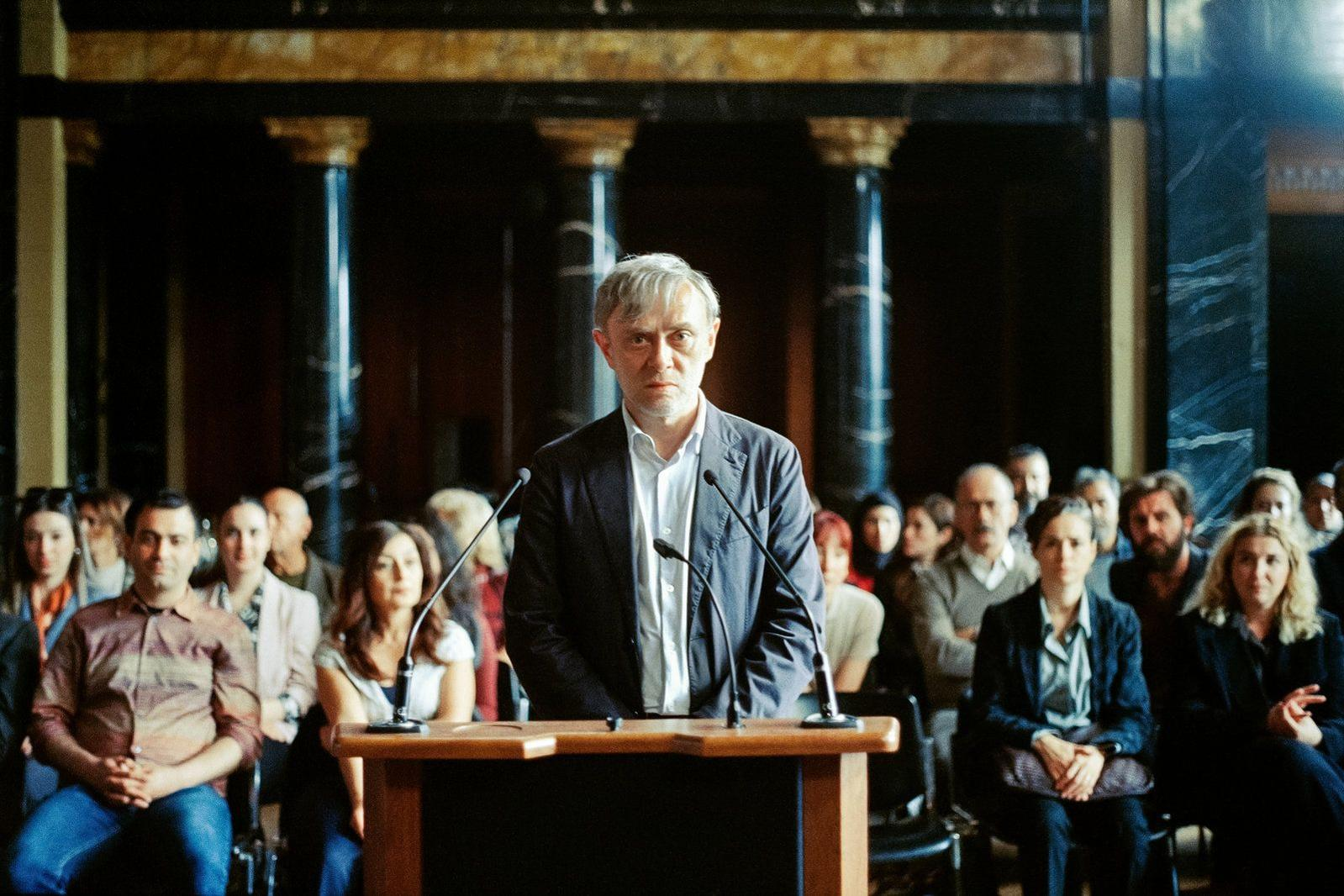 Ein Mann steht an einem Podium mit Mikrofonen vor einem Publikum in einem dunklen Raum mit Säulen.