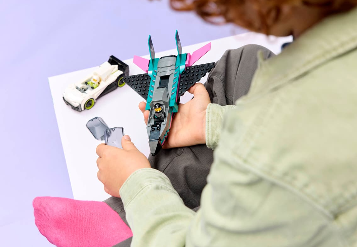 Kind spielt mit Lego-Spielzeug: ein graues Raketenschiff mit rosa Flügeln und ein weißes Auto.
