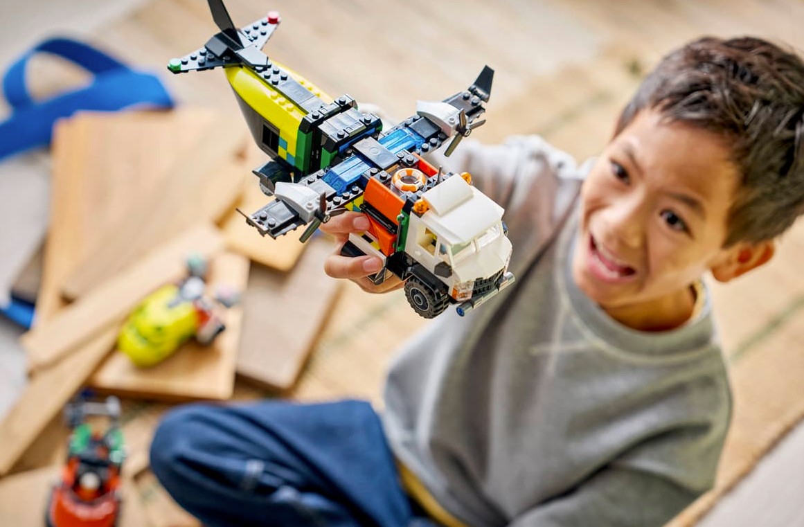 Ein Junge hält ein buntes Lego-Flugzeug mit LKW-Aufbau. Im Hintergrund liegen Holzbretter.