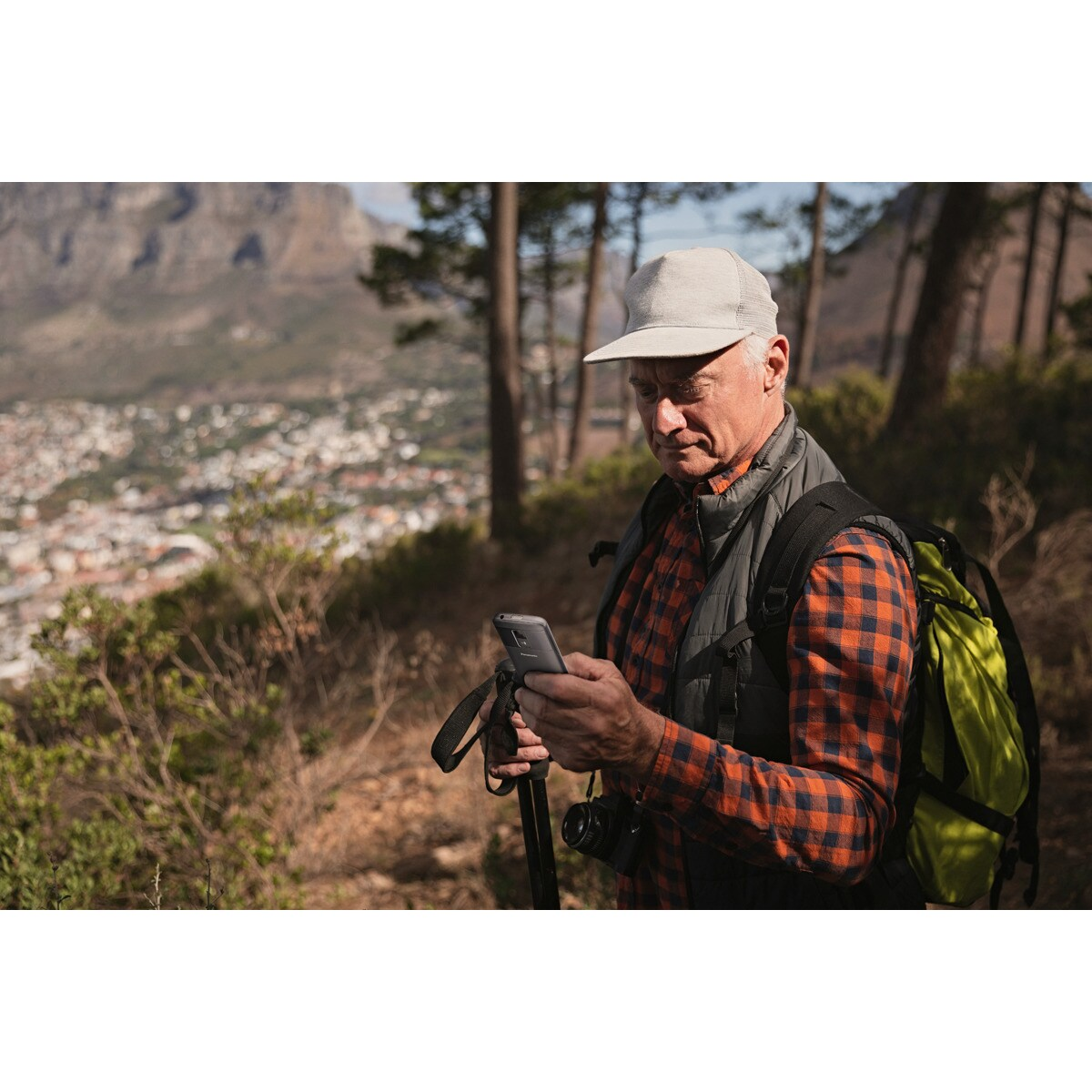 Hombre con gorra, chaleco y camisa a cuadros mira su teléfono, sosteniendo bastón. Mochila verde.