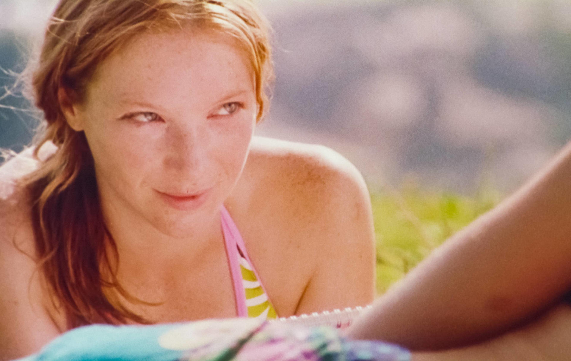 Ein junges Mädchen mit Sommersprossen und roten Haaren in einem Bikini liegt im Gras.