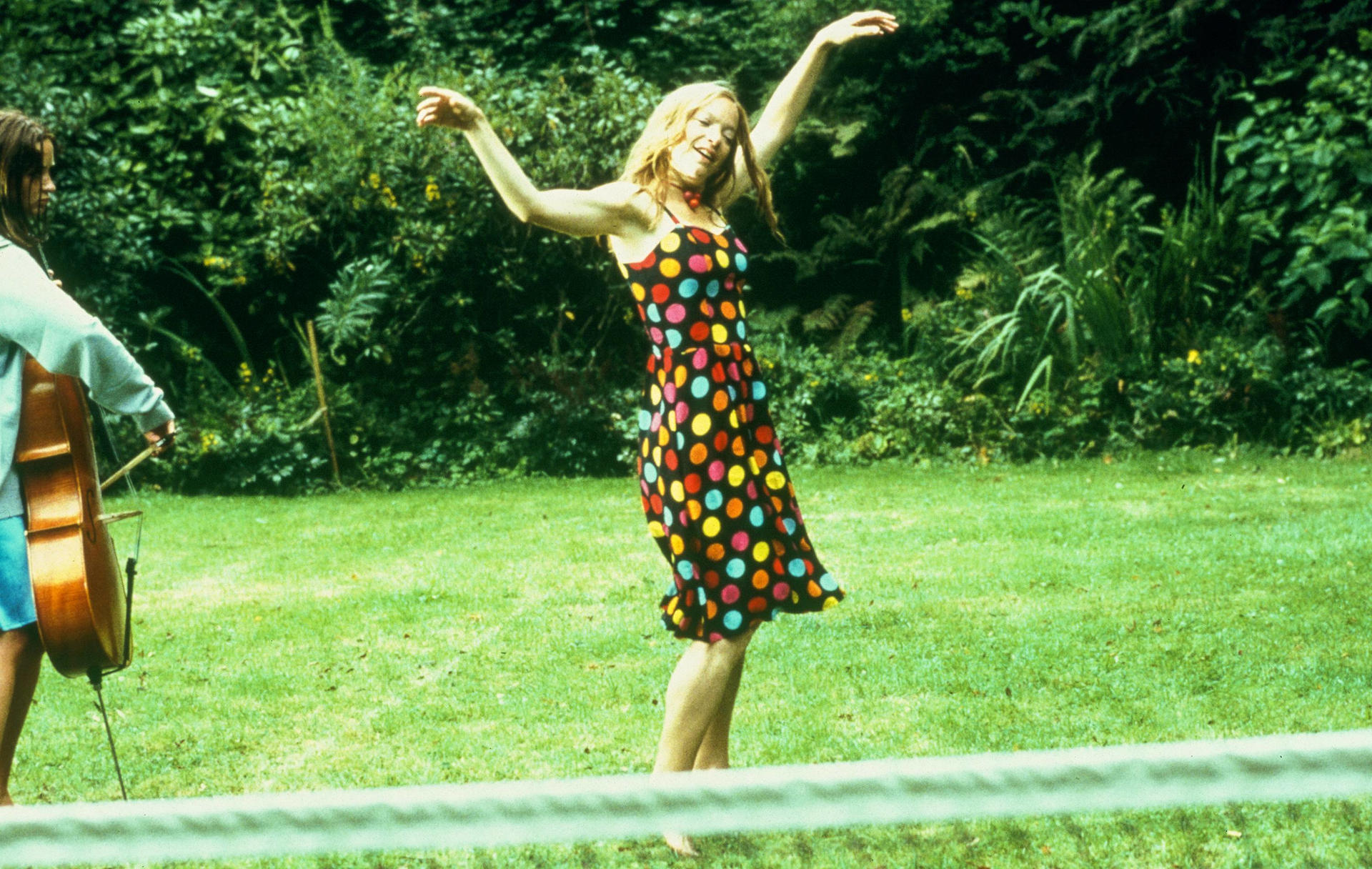 Eine Frau im gepunkteten Kleid tanzt im Garten, während in der Nähe jemand Cello spielt.
