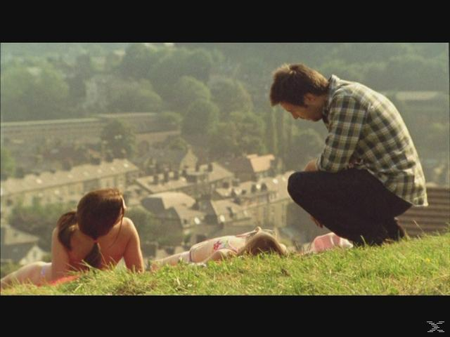 Ein Mann in kariertem Hemd hockt auf einem Hügel über einer Stadt. Zwei Personen liegen im Gras.