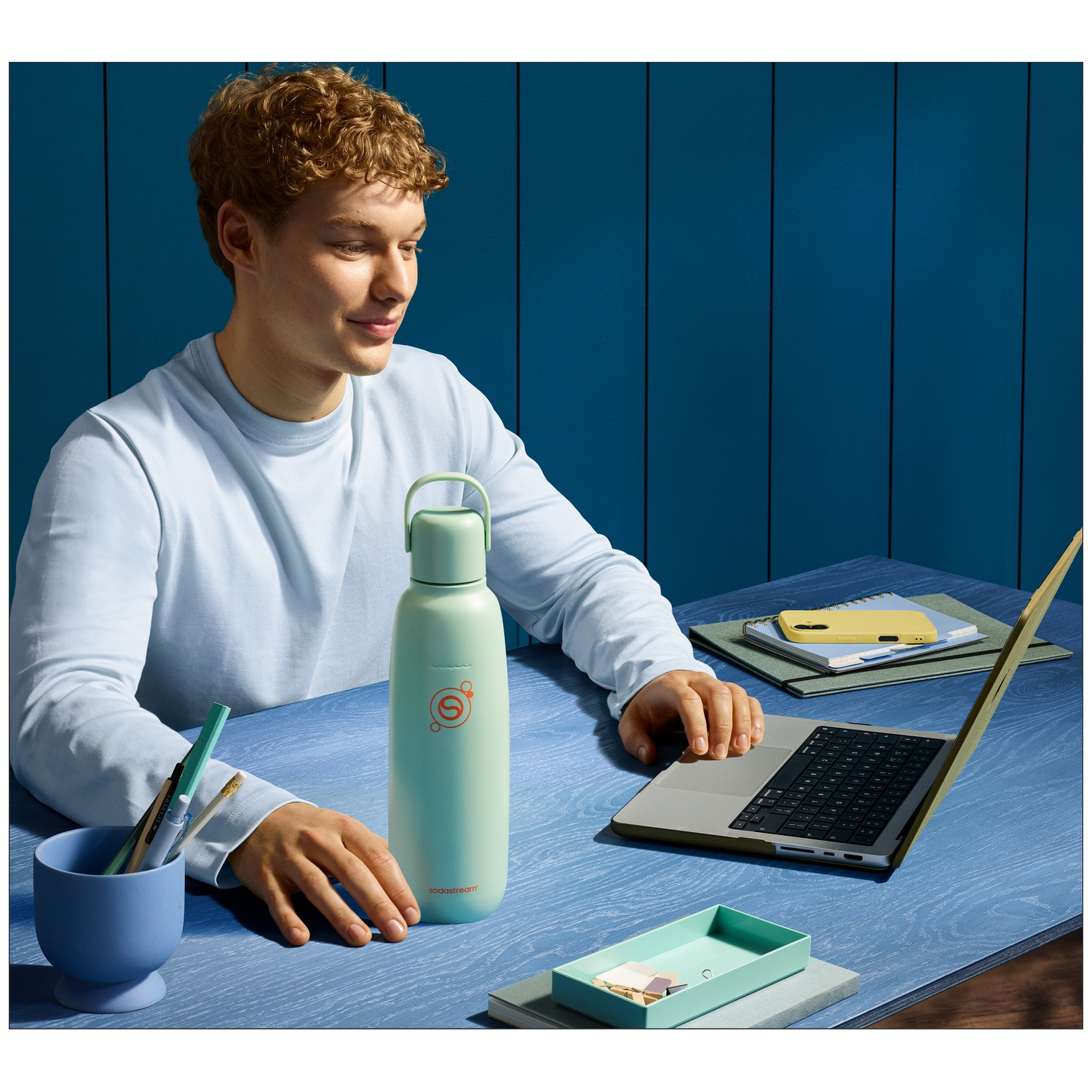 Homme à un bureau avec ordinateur portable, bouteille menthe, pot à stylos et plateau.