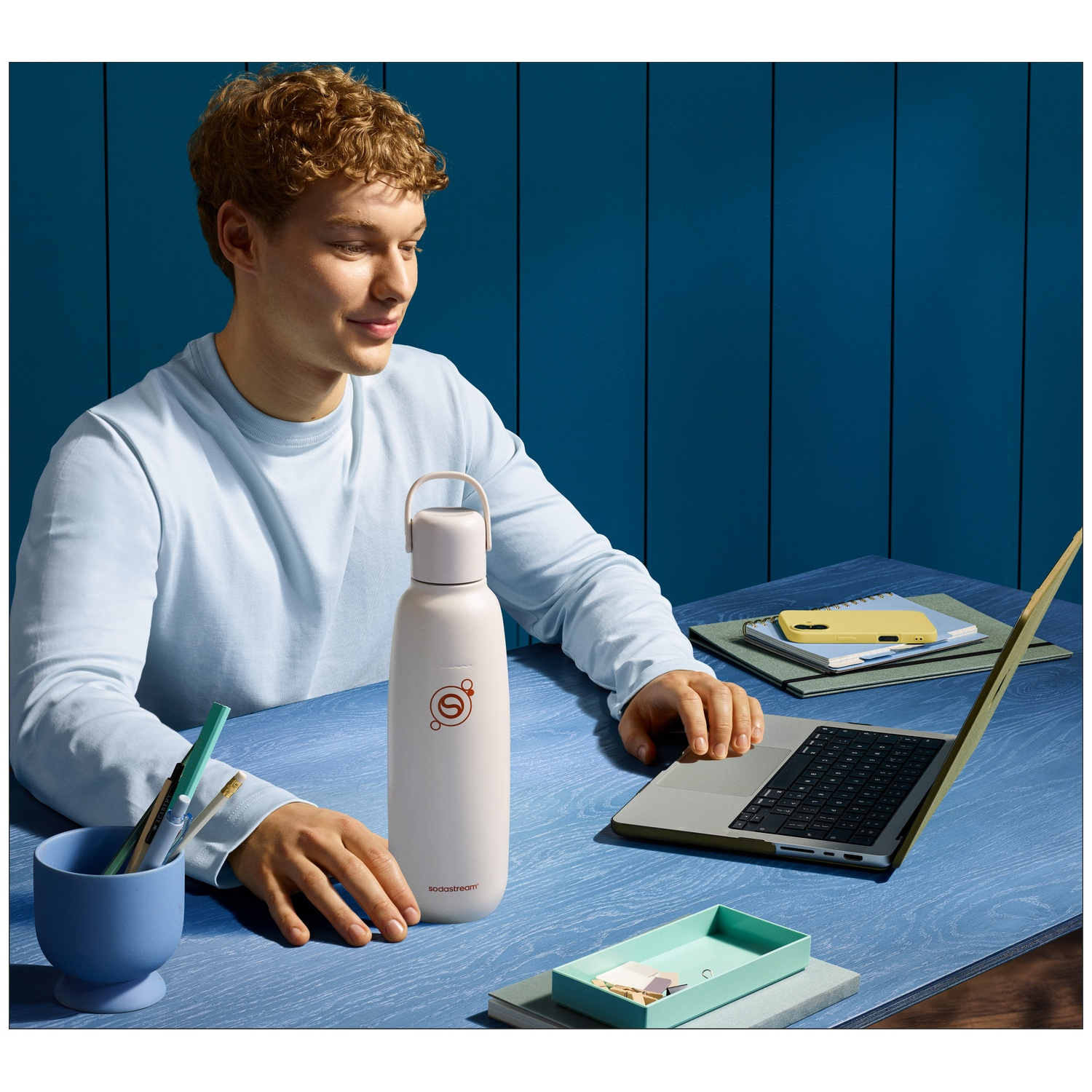 A young man sits at a blue table, looking at his laptop. A water bottle and pens are beside him.