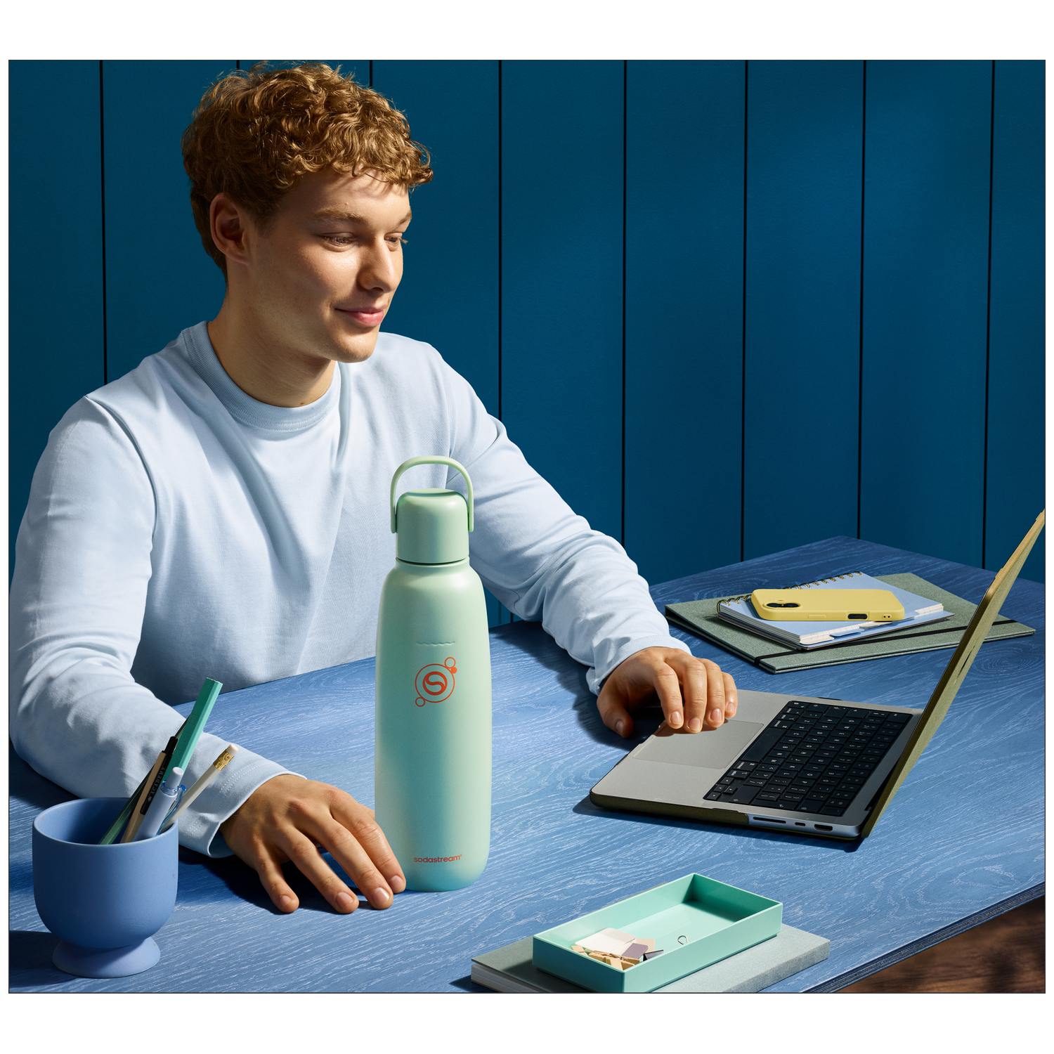 A young man sits at a desk with a water bottle and laptop nearby.