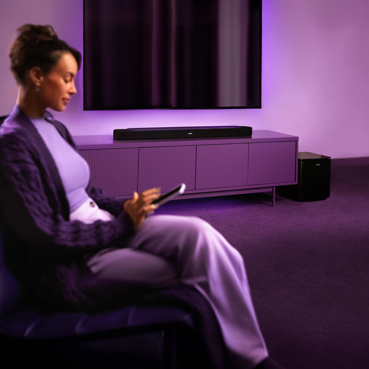 Eine Frau sitzt auf einem Sofa und schaut auf ein Tablet. Ein Fernseher, eine Soundbar und ein Subwoofer sind beleuchtet.