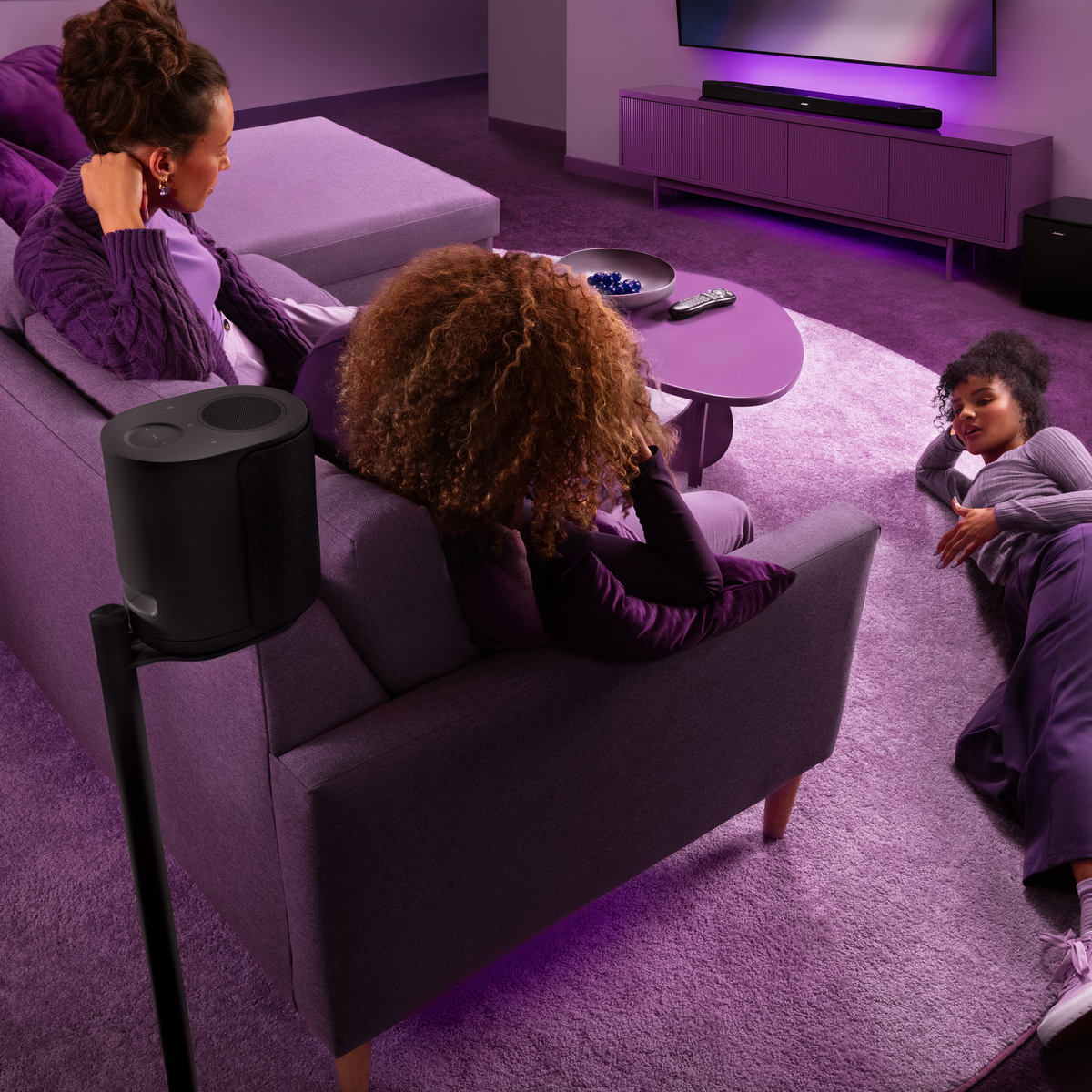 Drei Frauen entspannen sich in einem lila beleuchteten Wohnzimmer auf einem Sofa und dem Teppich.