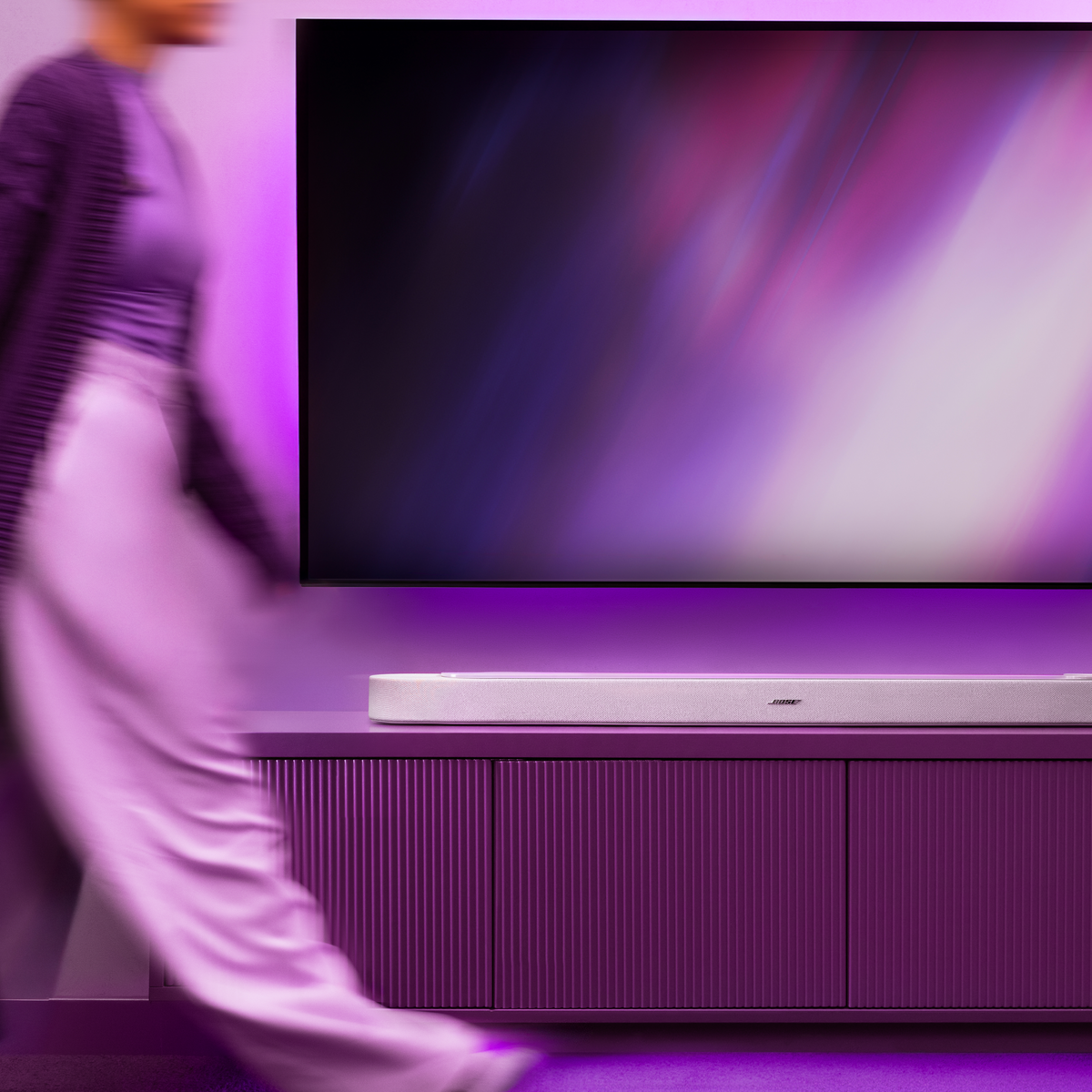 Eine Person in Lila geht an einem Fernsehgerät und einer Soundbar vorbei.