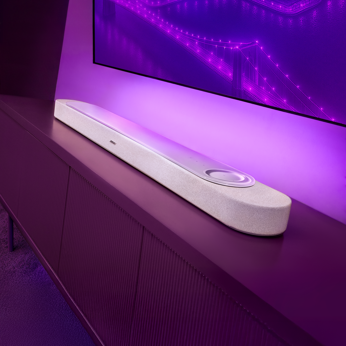 Ein weißer Soundbar steht auf einem lila Schrank vor einem lila beleuchteten Fernsehbildschirm.