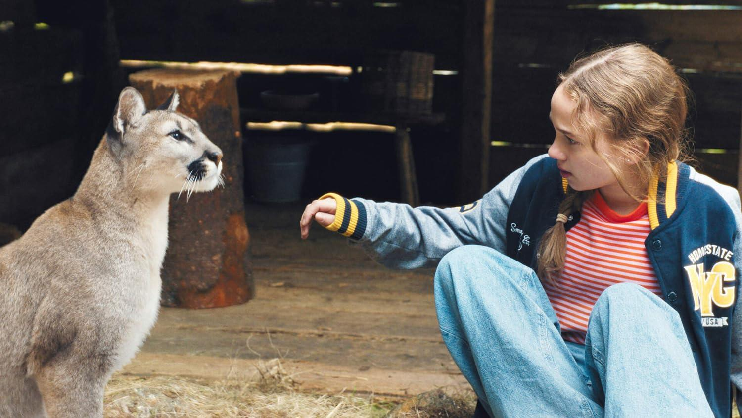 Ein Mädchen mit Zopf streckt die Hand zu einem Puma aus. Sie sitzen in einem rustikalen Holzschuppen.