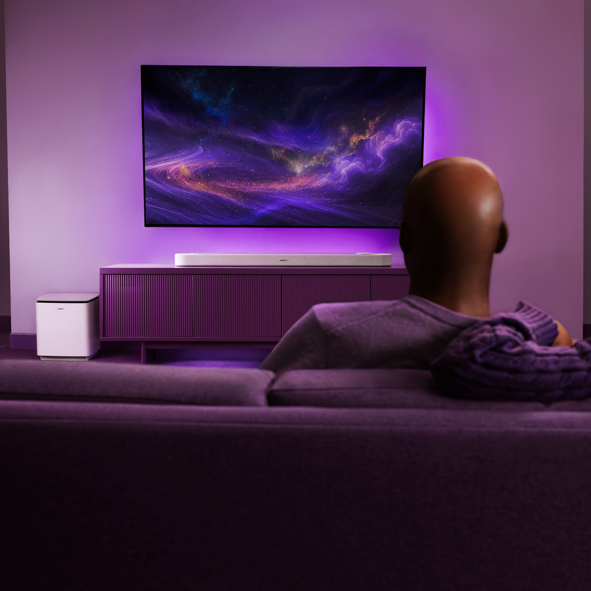 Mann sieht fern mit lila Umgebungslicht. Soundbar und Subwoofer sichtbar.