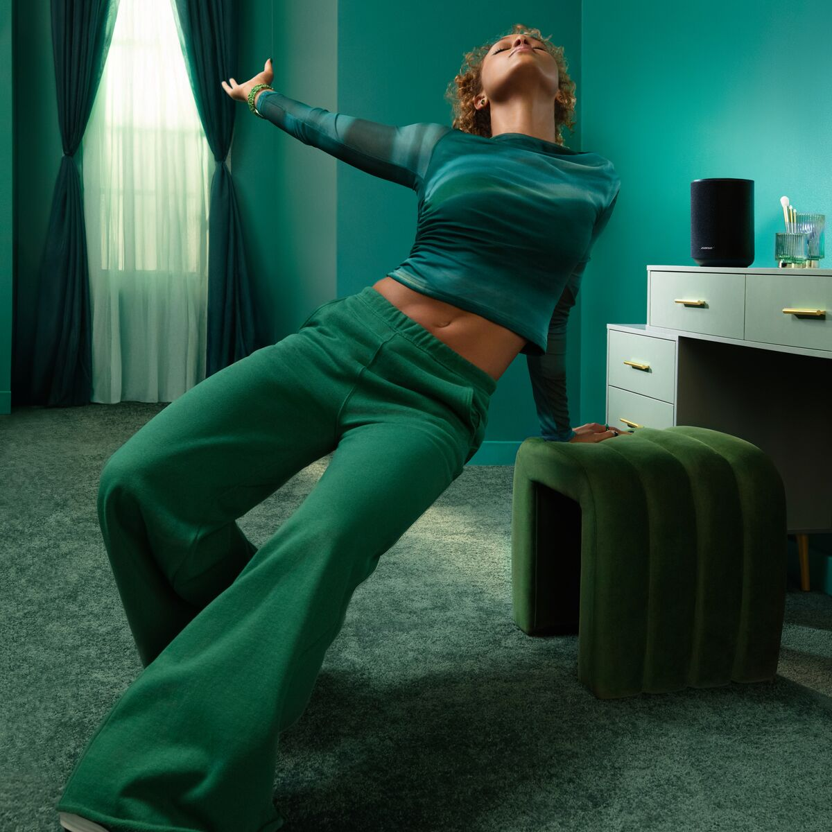 Eine Frau in grünen Klamotten lehnt sich auf einen grünen Hocker in einem teal-farbenen Raum.