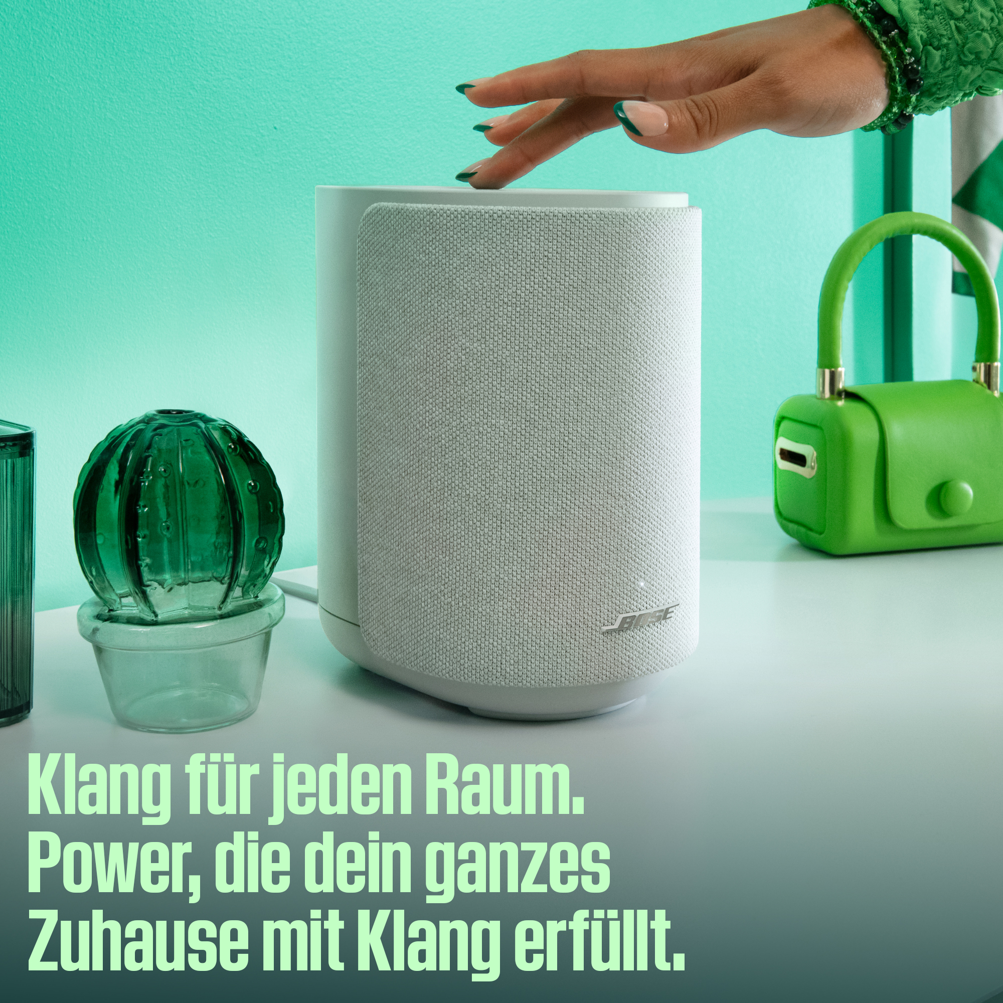 Eine Hand berührt einen weißen Lautsprecher neben einer grünen Tasche und Kaktusdeko.