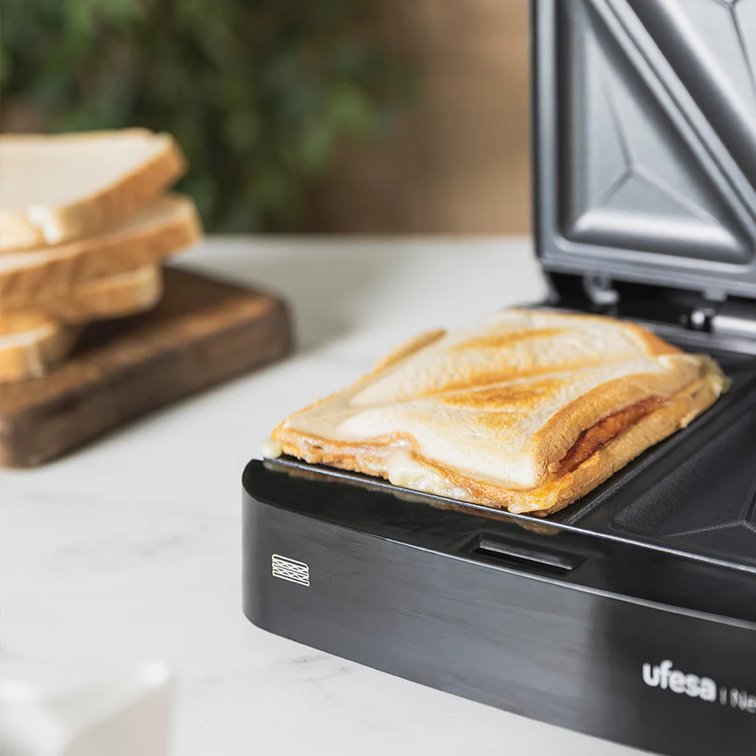 Un sándwich tostado con queso derretido en una sandwichera abierta. Pan tostado al fondo.