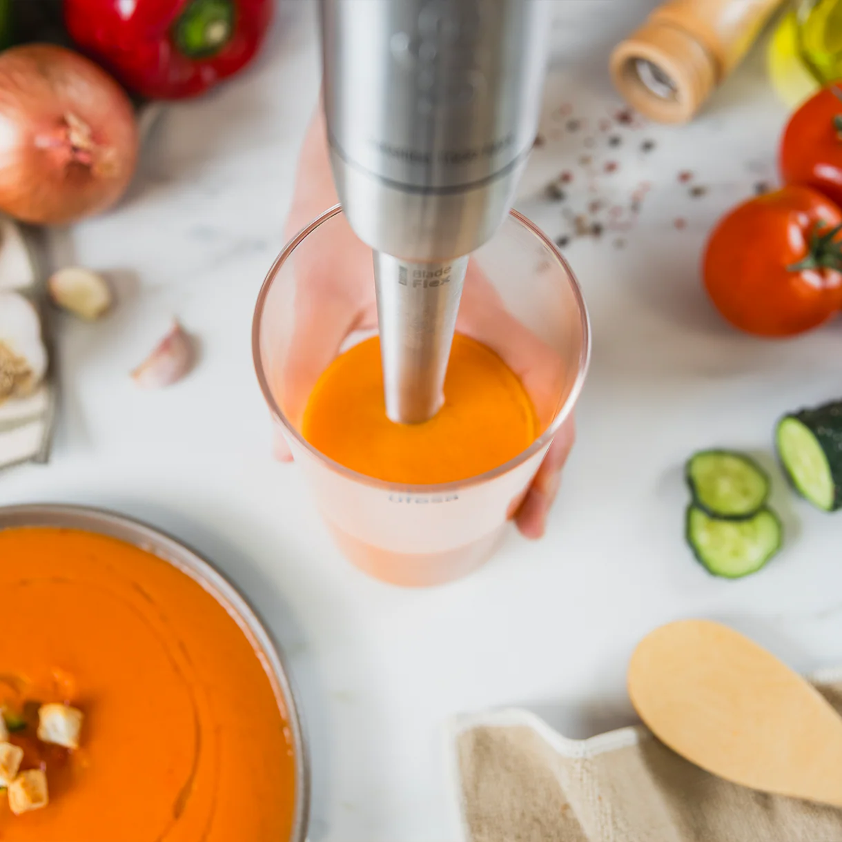 Una batidora de mano tritura líquido naranja en un vaso. Hay ingredientes cerca.