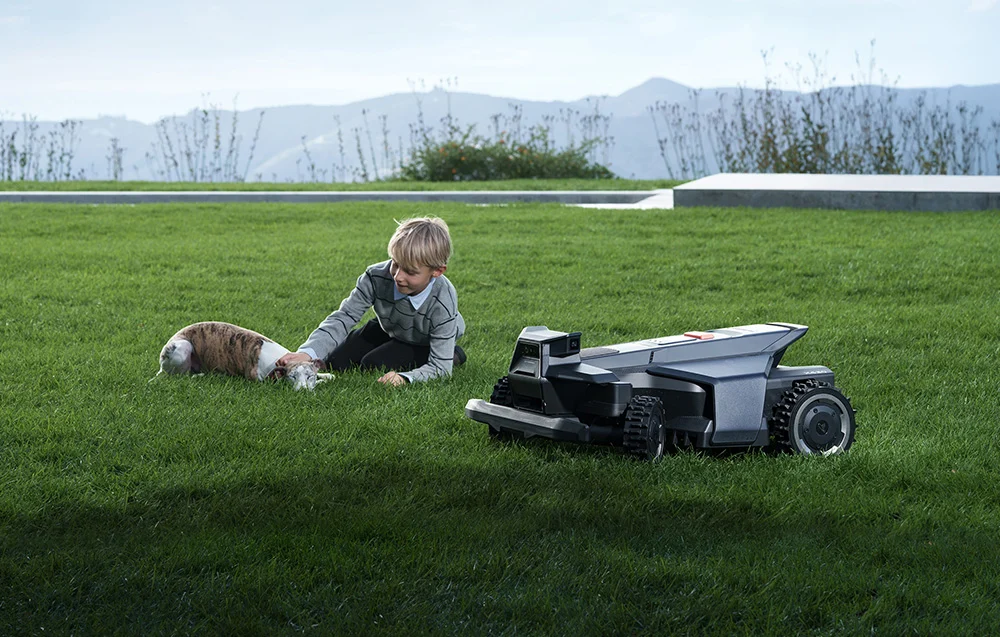 Egy fiú egy kutya simogat a pázsiton egy robotfűnyíró mellett.