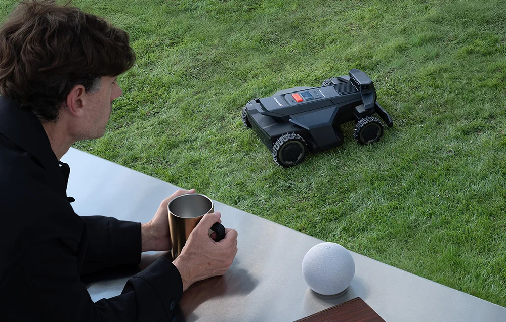 Egy férfi ül egy padon, bögréből iszik. Egy robotfűnyíró áll a füvön.