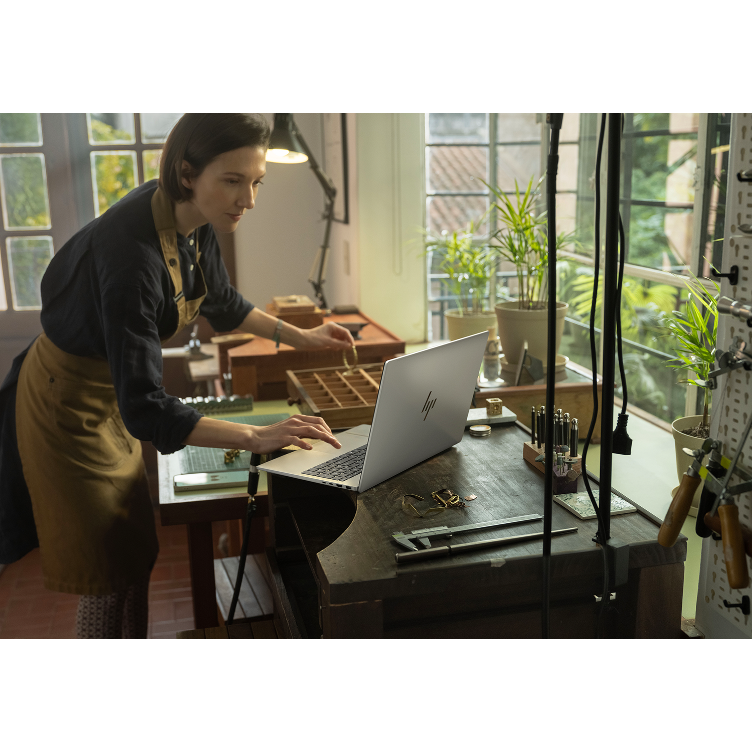 Een vrouw met schort werkt aan een laptop in een werkplaats met gereedschap.