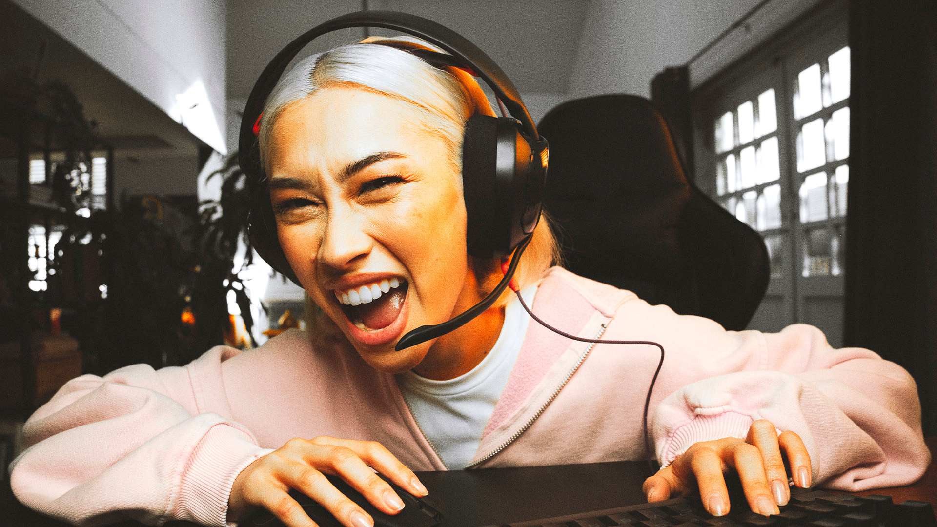 Eine Frau mit blonden Haaren und Headset lacht laut vor einem Computer.