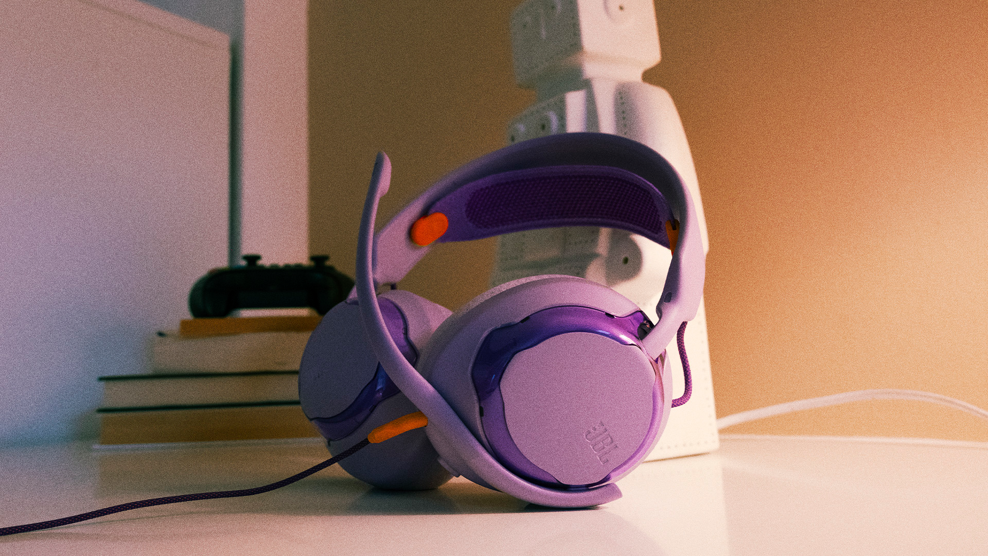Lila Kopfhörer mit Kabel stehen auf einem Tisch, hinter ihnen Bücher und ein Spielcontroller.
