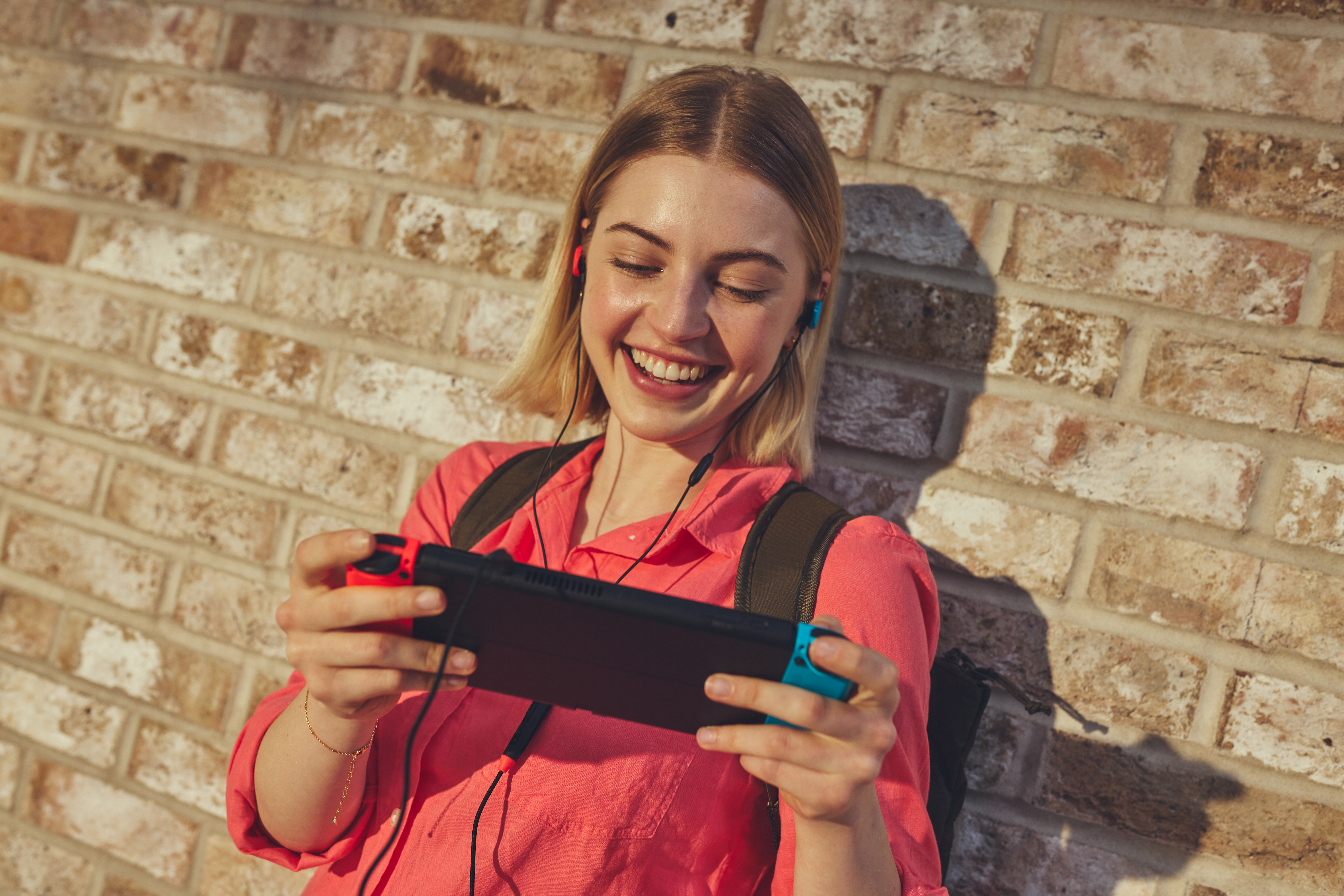 Jeune femme avec écouteurs joue à la Nintendo Switch devant un mur de briques.