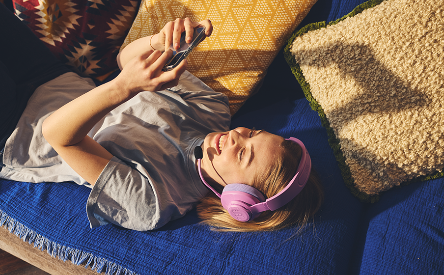 Una giovane persona con capelli biondi, che indossa una camicia azzurra e cuffie rosa, sdraiata su una coperta blu e con in mano un telefono.