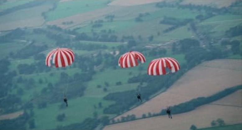 Drei Fallschirmspringer mit rot-weißen Fallschirmen über grünen Feldern.