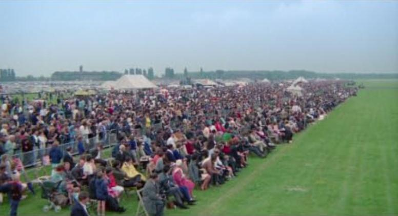 Große Menschenmenge sitzt auf Stühlen auf einem Feld; Zelte und Bäume im Hintergrund.