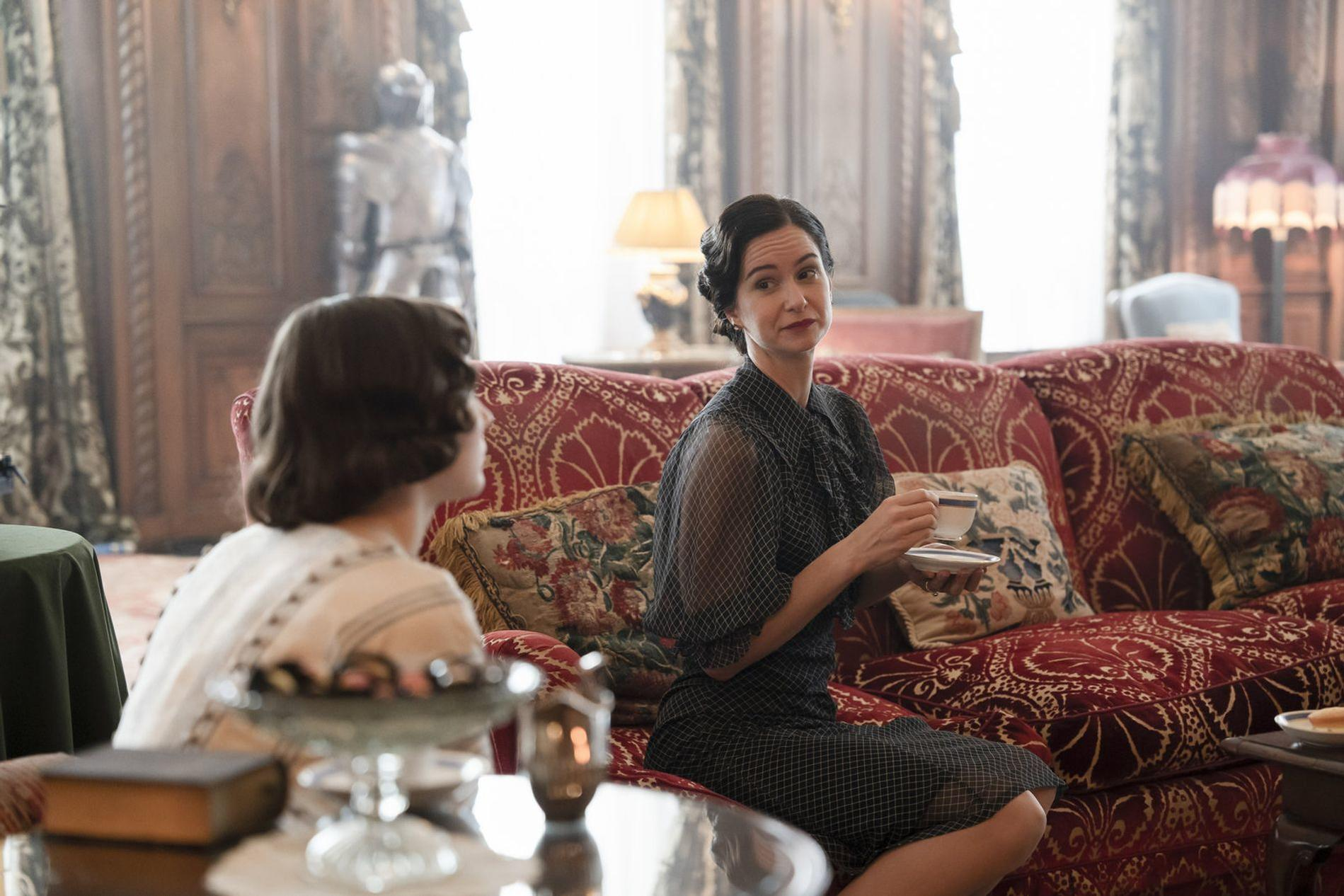 Zwei Frauen sitzen auf einem roten Sofa in einem opulenten Raum. Eine hält eine Tasse.