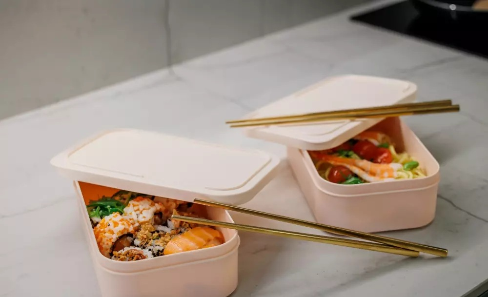 Dwa różowe pudełka na lunch z sushi i makaronem, obok pałeczki.