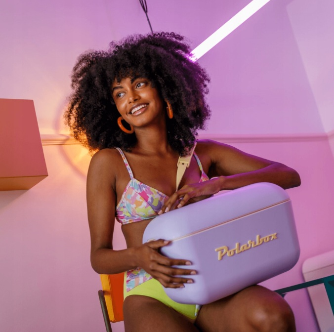 Kobieta z bujnymi włosami trzyma lawendową Polarbox, nosi bikini. Neonowe światła.