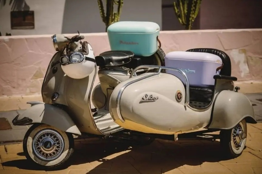 Kremowa Vespa z wózkiem bocznym i dwoma lodówkami, jedną jasnoniebieską, drugą fioletową.